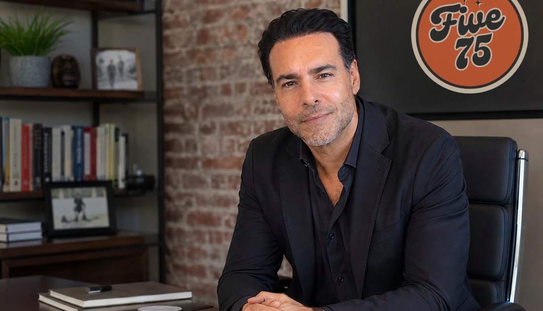 Joe Luna, founder of FIVE75, seated at his desk with the FIVE75 retro badge on the brick wall behind him